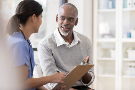 A cheerful senior man sits next to an unrecognizable female doctor in her office.  She holds a clipboard as he asks questions regarding his diagnosis.