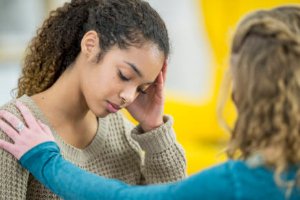 Two young women are indoors in a bedroom. They are wearing casual clothing. One woman looks sad, and she is being comforted by her friend.
